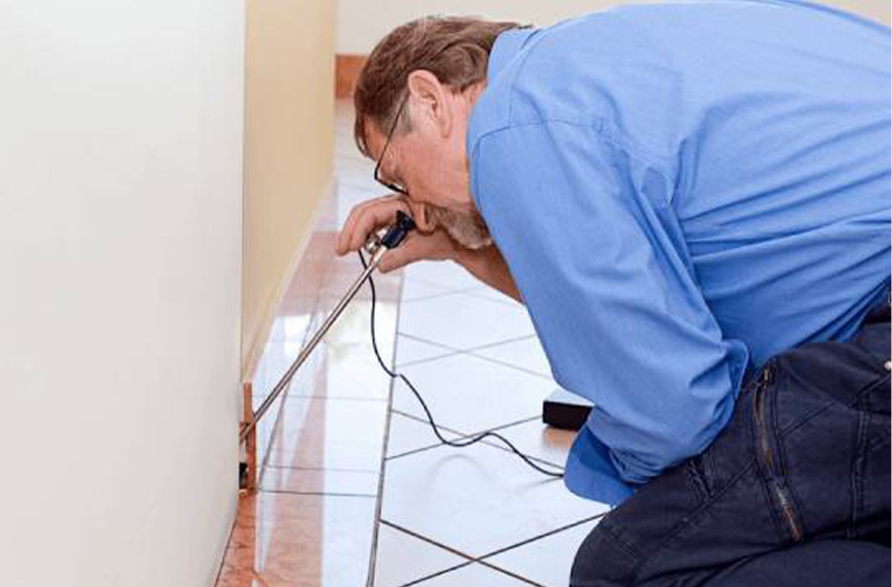 Inspector kneeling on a tiled floor testing a skirting board with a moisture meter probe