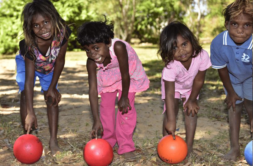 Young children smiling as they bend over red and orange balls on a dirt playing field