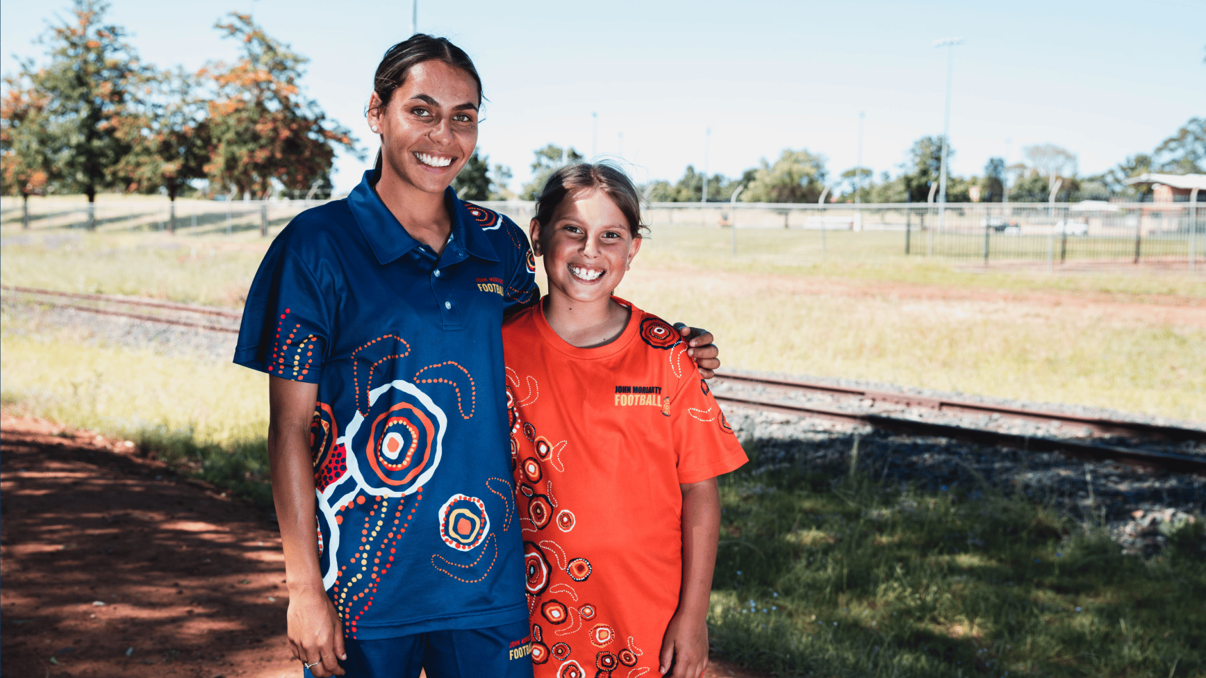 Young woman and a girl in Aboriginal-design football shirts smiling beside a railway line
