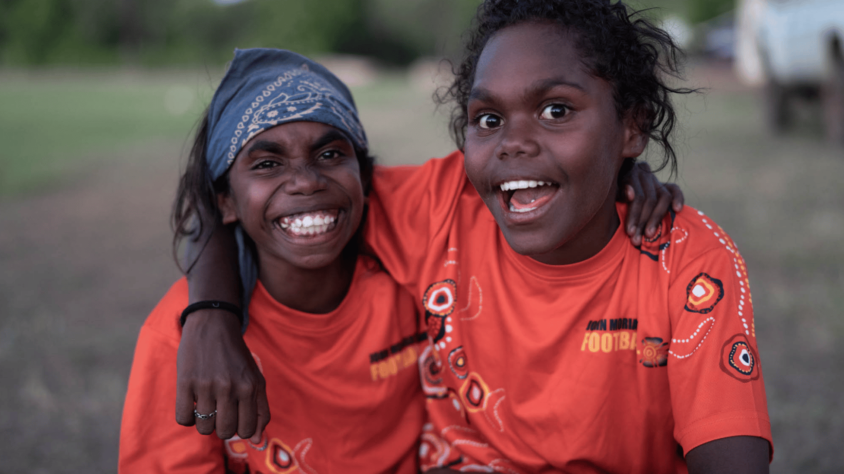 Two smiling girls in orange John Moriarty Football shirts with arms around each other