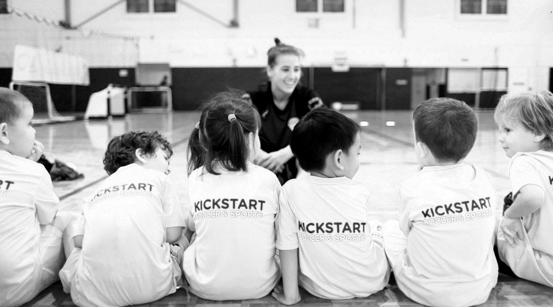 Sports Kickstart coach talking with children in Kickstart shirts on a gym floor