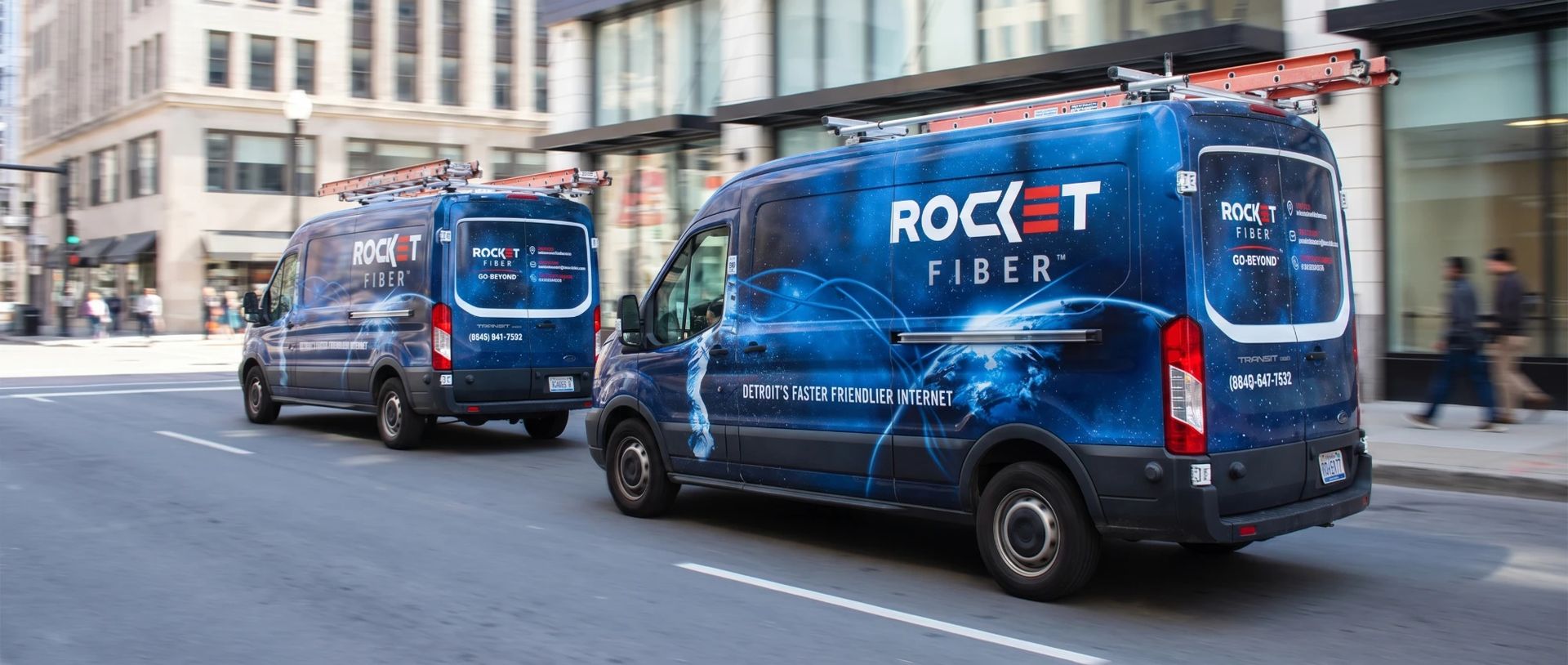 Two Rocket Fiber vans with roof ladders driving through a downtown street