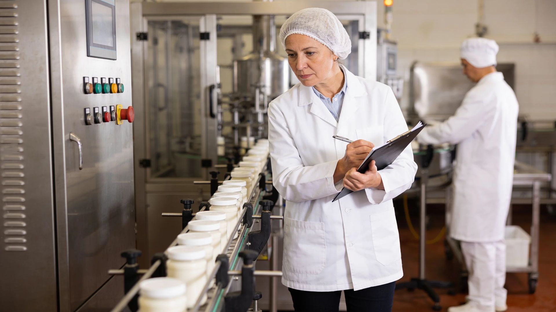 Worker in a lab coat writing on a clipboard beside a filling line